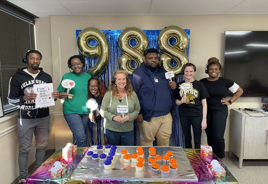 Santé- The Helpers You Dont Always See - Santé Group of coworkers posing in an office with a table of orange and purple cupcakes and a blue foil backdrop behind them; large gold '98' balloons in the background.
