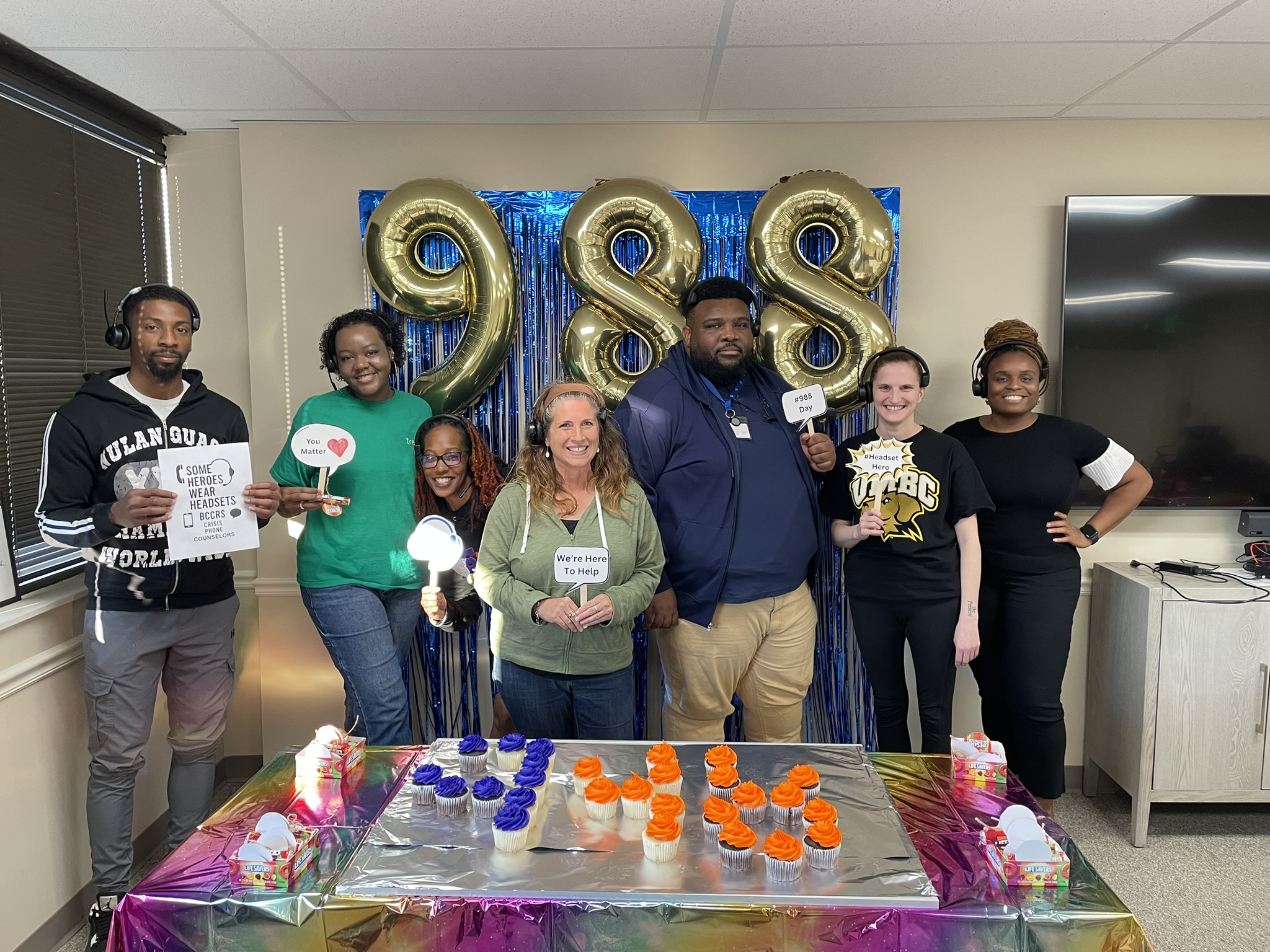 Group of coworkers posing in an office with a table of orange and purple cupcakes and a blue foil backdrop behind them; large gold '98' balloons in the background.