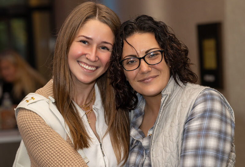 The-Hidden-Factors-Affecting-Mental-Health-Why-Crisis-Services-Must-Address-More-Than-the-Crisis-e1783440314403-1 - Santé Two women smiling and posing together indoors, one with glasses and plaid shirt, the other with long blonde hair and a white vest flecked with knit sleeves.